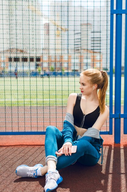 Una giovane ragazza in una tuta sportiva blu con un top nero è seduta vicino al recinto dello stadio. Sta ascoltando la musica con le cuffie. Ha una figura attraente.