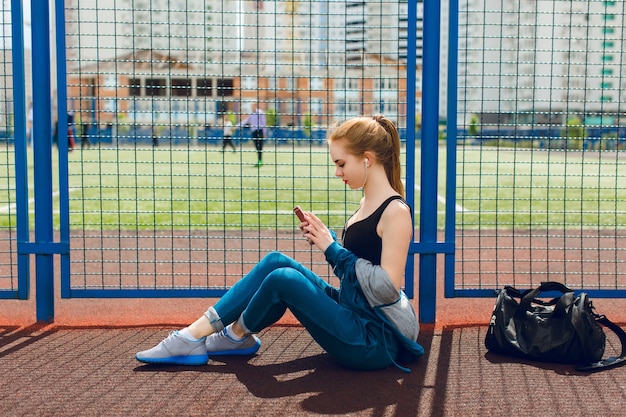 Una giovane ragazza in una tuta sportiva blu con un top nero è seduta vicino al recinto dello stadio. Sta ascoltando la musica con le cuffie.