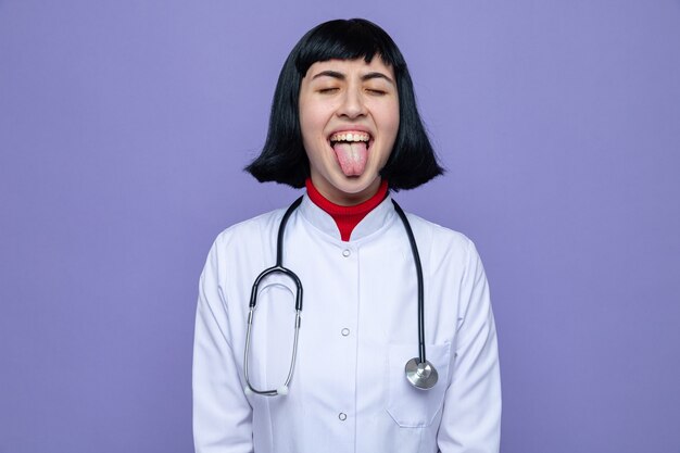 Una giovane ragazza piuttosto caucasica divertente in uniforme da medico con stetoscopio sporge la lingua in piedi con gli occhi chiusi