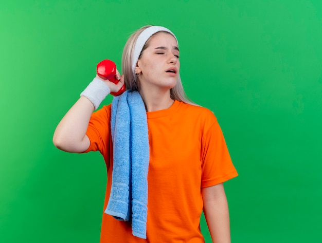 Una giovane ragazza sportiva caucasica scontenta con bretelle che indossa fascia e braccialetti con un asciugamano sulla spalla tiene il manubrio