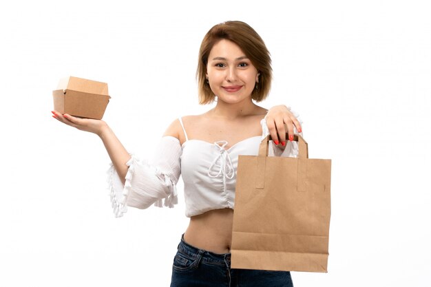 Una giovane signora attraente di vista frontale in camicia bianca e blue jeans che tengono pacchetto marrone e piccola scatola felici sul bianco