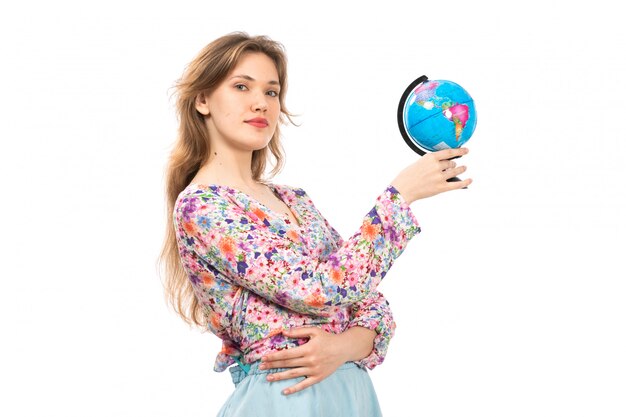 Una giovane signora attraente di vista frontale in camicia colorata fiore progettato e gonna blu che tiene piccolo globo sul bianco