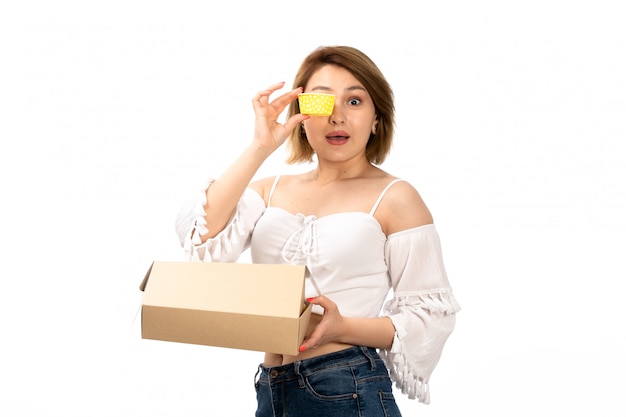 Una giovane signora attraente di vista frontale in camicia e blue jeans bianche che ottengono piccolo pacchetto che lo apre tenendo presente giallo sul bianco