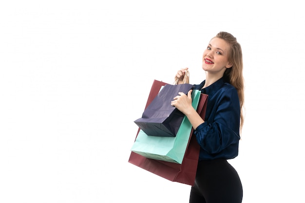 Una giovane signora attraente di vista frontale in pantaloni neri della blusa blu che posano sorridere dei pacchetti di acquisto della tenuta felice sui vestiti bianchi di modo del fondo bianco