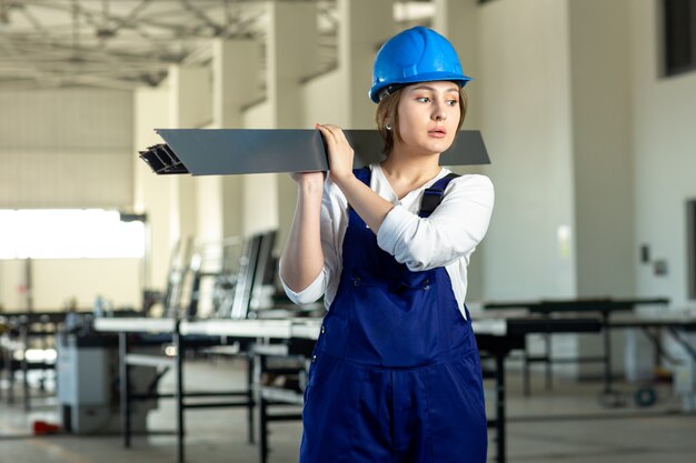 Una giovane signora attraente di vista frontale in vestito blu della costruzione e casco che lavorano tenendo dettaglio metallico pesante durante la costruzione di architettura di giorno delle costruzioni