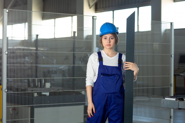 Una giovane signora attraente di vista frontale in vestito blu della costruzione e casco che lavorano tenendo dettaglio metallico pesante durante la costruzione di architettura di giorno delle costruzioni