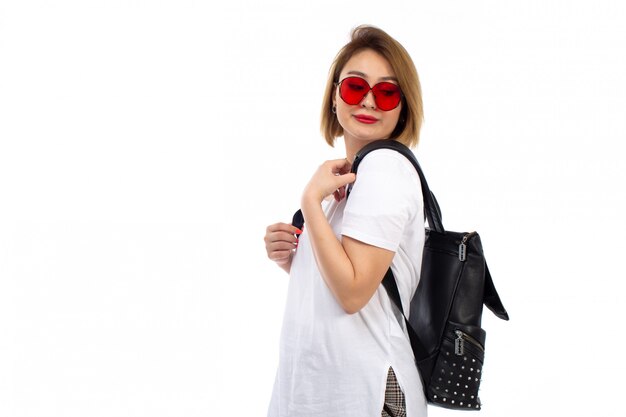 Una giovane signora di vista frontale in borsa nera degli occhiali da sole rossi della maglietta bianca che sorride sul bianco