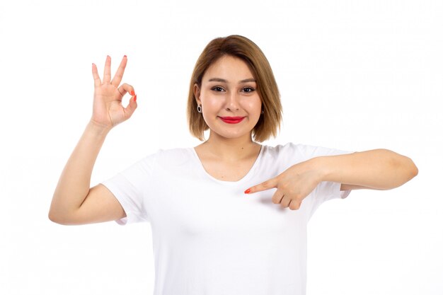 Una giovane signora di vista frontale in camicia bianca che posa sorridere mostrando segno giusto sul bianco