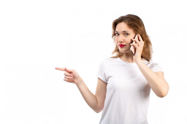 Una giovane signora di vista frontale in camicia bianca e jeans moderni neri che posano parlare sul telefono sul bianco