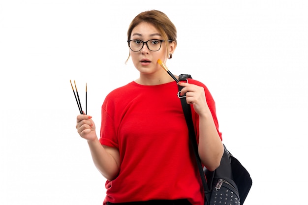 Una giovane studentessa di vista frontale in jeans neri della maglietta rossa che tengono i pennelli sul bianco