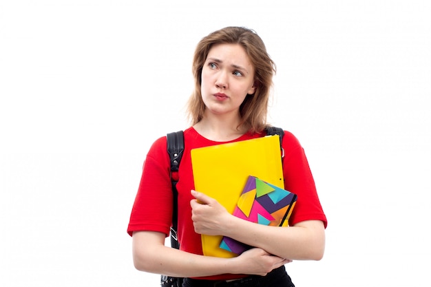 Una giovane studentessa di vista frontale negli archivi rossi dei quaderni della tenuta della borsa del nero della camicia che pensa sul bianco