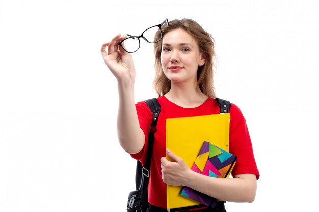 Una giovane studentessa di vista frontale negli archivi rossi dei quaderni della tenuta della borsa del nero della camicia che sorride sul bianco