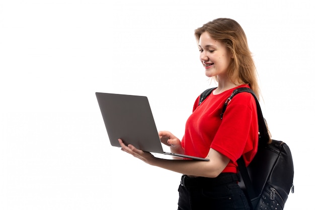 Una giovane studentessa di vista frontale nella borsa rossa del nero della camicia facendo uso del computer portatile sul bianco