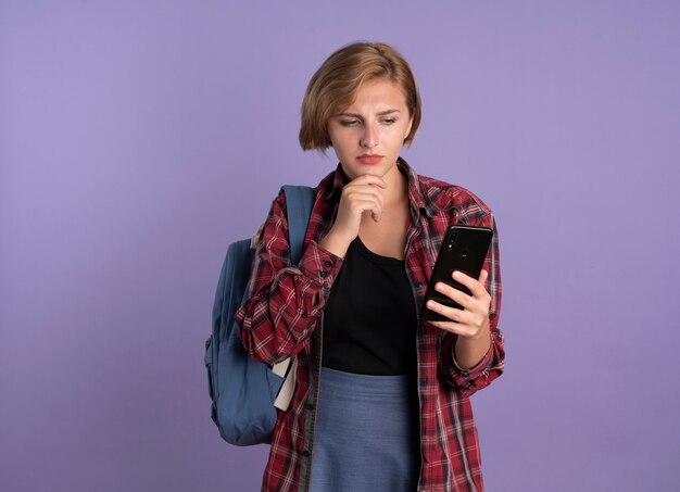 Una giovane studentessa slava confusa che indossa uno zaino mette la mano sul mento e guarda il telefono