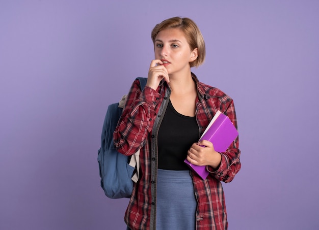Una giovane studentessa slava confusa che indossa uno zaino morde un chiodo tiene un libro e un taccuino