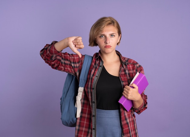 Una giovane studentessa slava scontenta che indossa lo zaino pollice in giù tiene libro e quaderno