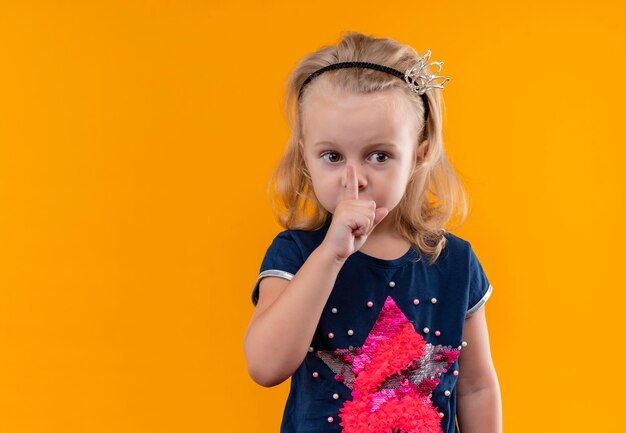 Una graziosa bambina che indossa una maglietta blu navy nella fascia della corona che mostra il gesto di shh con il dito indice sulla bocca e guardando il lato su una parete arancione