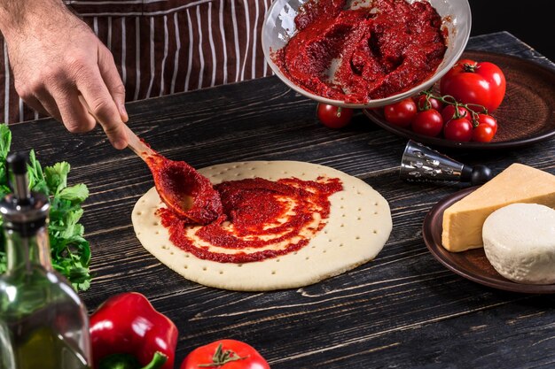 Una mano maschio che sparge la passata di pomodoro su una base di pizza con il cucchiaio su un vecchio fondo di legno. Concetto di cucina. Avvicinamento