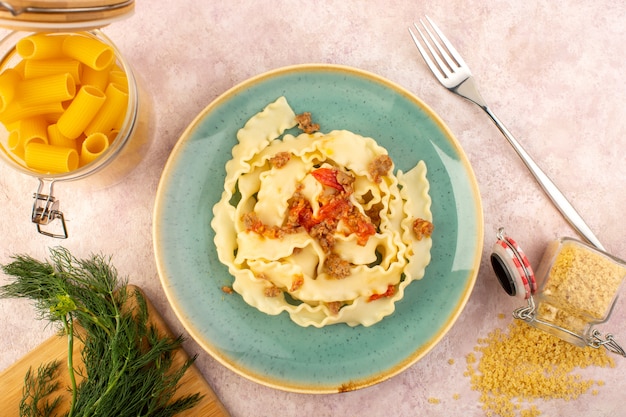 Una pasta italiana con vista dall'alto cucinata gustosa salata all'interno di un piatto verde rotondo con verdure e pasta cruda sulla scrivania rosa