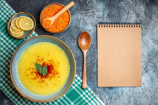 Una pentola blu con una gustosa zuppa servita con menta e pepe accanto a un cucchiaio di legno di limone tritato e un quaderno a spirale di piselli gialli su sfondo blu