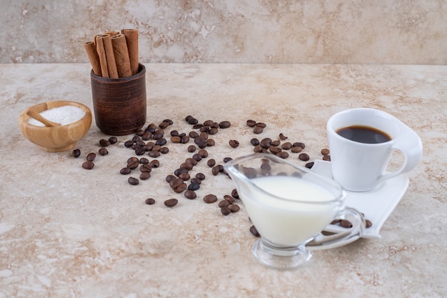 Una piccola ciotola di zucchero e bastoncini di cannella in una tazza di legno accanto a chicchi di caffè sparsi, che serve un bicchiere di latte e una tazza di caffè