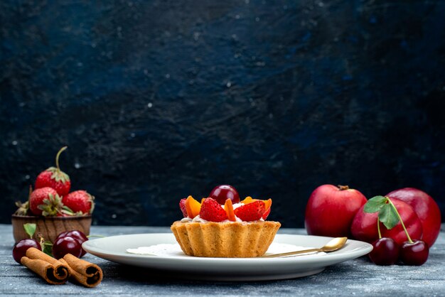 Una piccola torta deliziosa di vista frontale con crema e frutta all'interno del piatto bianco insieme a frutta fresca sul biscotto della torta di frutta della scrivania grigio-blu