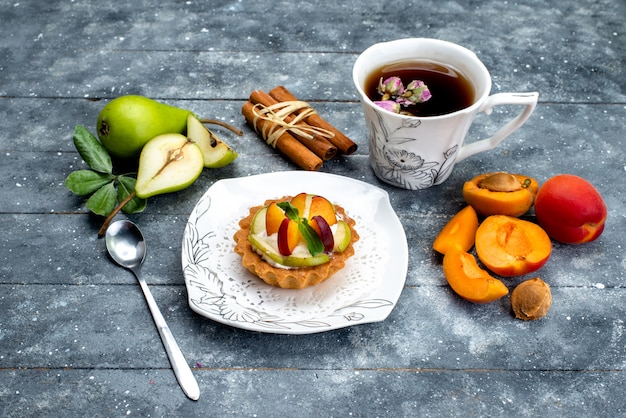 Una piccola torta deliziosa vista dall'alto con crema e frutta a fette all'interno del piatto con tè sul biscotto torta scrivania grigia