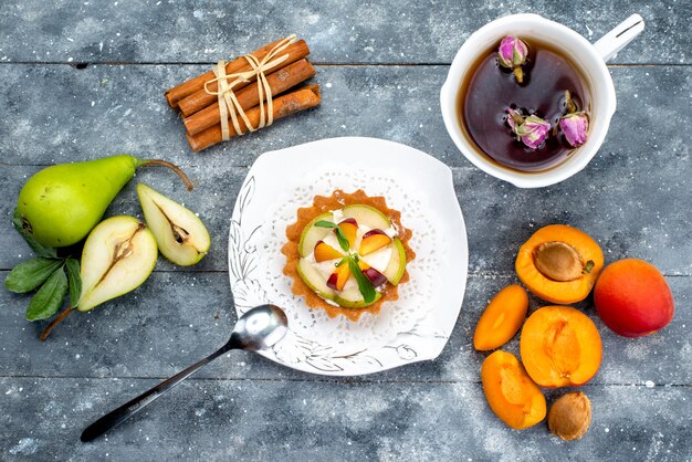 Una piccola torta deliziosa vista dall'alto con crema e frutta a fette all'interno del piatto con tè sul tè biscotto torta scrivania grigia