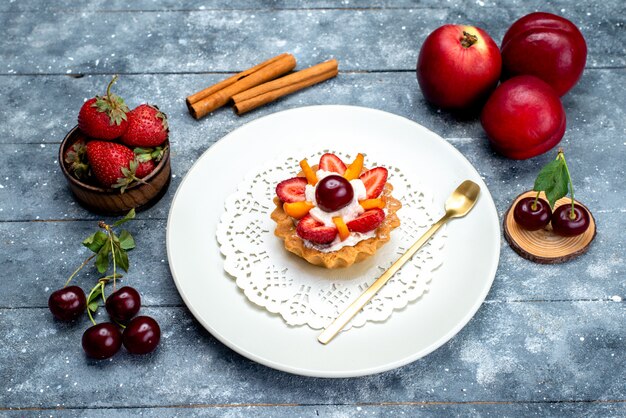 Una piccola torta deliziosa vista dall'alto con crema e frutta all'interno del piatto bianco insieme a frutta fresca sul biscotto alla frutta da scrivania grigio-blu