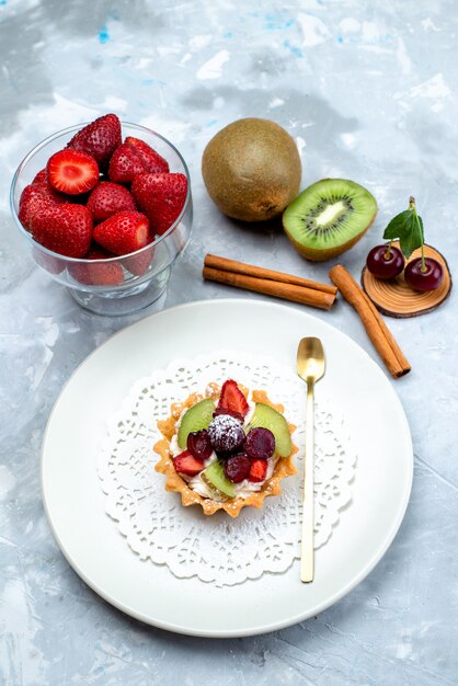 Una piccola torta deliziosa vista dall'alto con crema e frutta all'interno del piatto bianco insieme alla cannella di frutta fresca sulla torta di frutta da scrivania grigio-blu