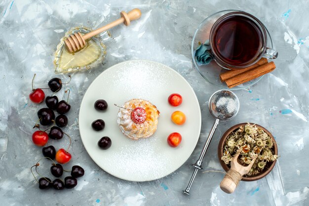 Una piccola torta di vista superiore all'interno del piatto bianco con il tè delle ciliege e la cannella sullo zucchero leggero del biscotto della torta dello scrittorio