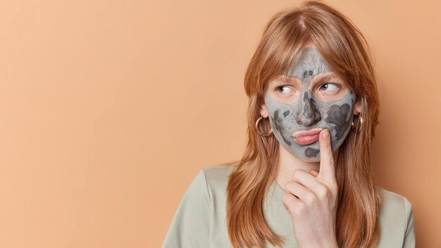 Una ragazza adolescente seria e pensierosa con i capelli rossi distoglie lo sguardo e pensa a come migliorare le condizioni della pelle applica una maschera di argilla sul viso isolato su sfondo beige copia spazio per la tua promozione Concetto di bellezza