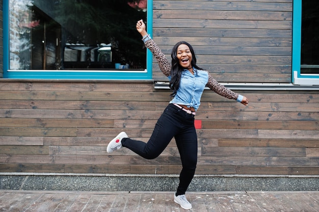 Una ragazza afroamericana hipster che indossa una camicia di jeans con maniche di leopardo salta in strada contro una casa di legno con finestre