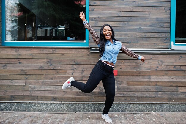 Una ragazza afroamericana hipster che indossa una camicia di jeans con maniche di leopardo salta in strada contro una casa di legno con finestre