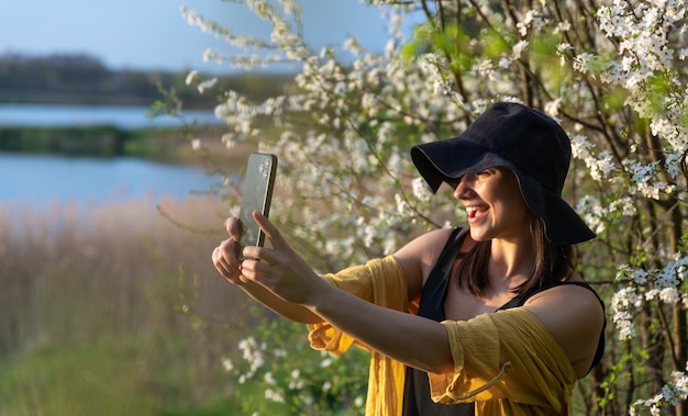 Una ragazza alla moda con un cappello fa un selfie al tramonto vicino agli alberi in fiore nella foresta