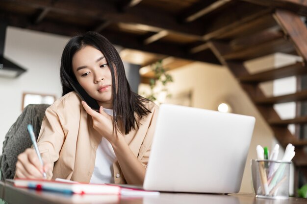 Una ragazza asiatica occupata risponde alla telefonata prendendo appunti scrivendo sul taccuino lavorando da casa con un pc portatile