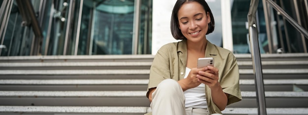 Una ragazza asiatica sorridente si siede sulle scale vicino all'ingresso dell'edificio utilizzando l'app per telefono cellulare giovane donna felice