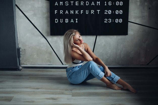 Una ragazza attraente con i capelli biondi in jeans si siede sul pavimento accanto al tabellone con un programma e pensa a qualcosa