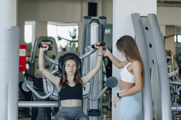 Una ragazza attraente in palestra modella un gruppo superiore di muscoli, braccia e spalle sotto la supervisione di un allenatore.