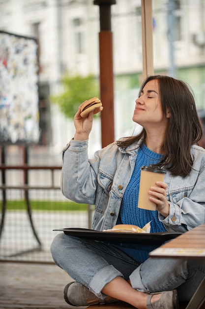Una ragazza attraente in stile casual mangia un hamburger con un caffè seduto sulla terrazza estiva