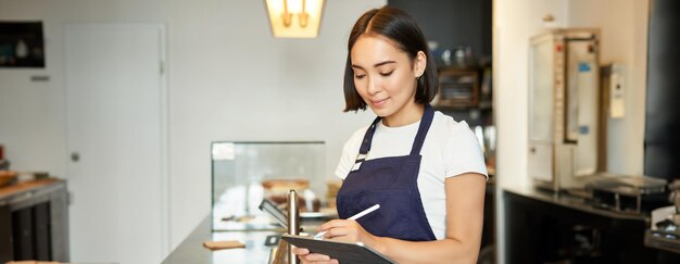 Una ragazza barista carina sorridente, una donna coreana che lavora in un caffè usando un tablet per elaborare l'ordine di caffè