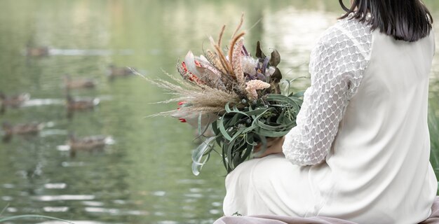 Una ragazza bruna in abito bianco si siede in riva al fiume con un mazzo di fiori esotici, sfondo sfocato, vista posteriore.