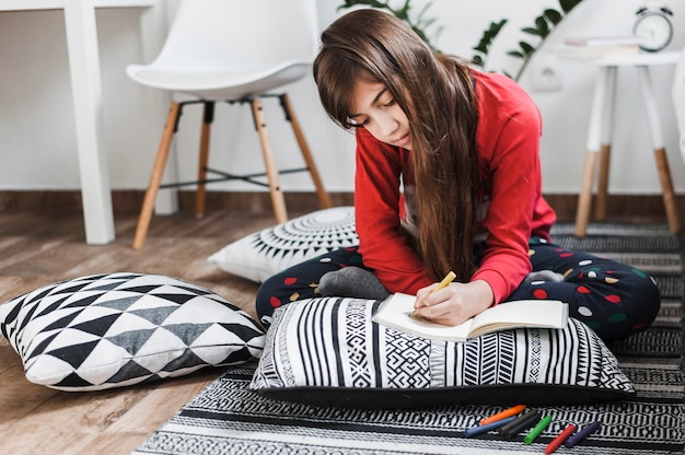 Una ragazza che si siede sul tappeto di disegno con i pastelli colorati sul notebook