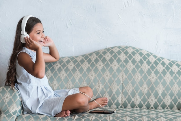 Una ragazza che si siede sulla musica d&#39;ascolto del sofà sulla cuffia allegata al telefono cellulare