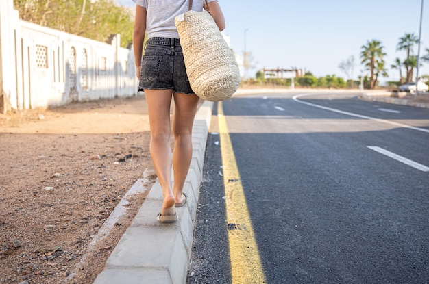 Una ragazza con le gambe abbronzate cammina lungo il marciapiede lungo la strada con la borsa.