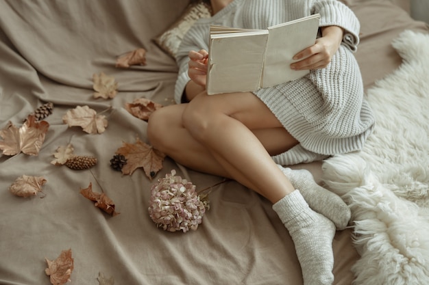 Una ragazza con un comodo maglione lavorato a maglia e calzini caldi giace a letto con un libro tra le foglie autunnali.