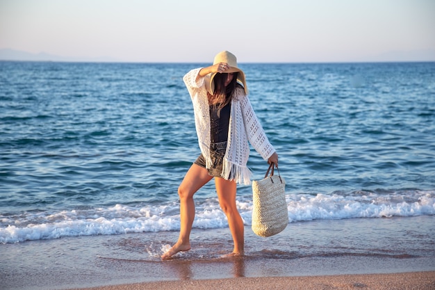 Una ragazza con un grande cappello con una borsa di vimini cammina in riva al mare. Concetto di vacanza estiva.