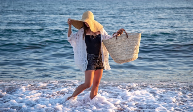 Una ragazza con un grande cappello e una borsa di vimini cammina lungo la costa del mare. Concetto di vacanza estiva.