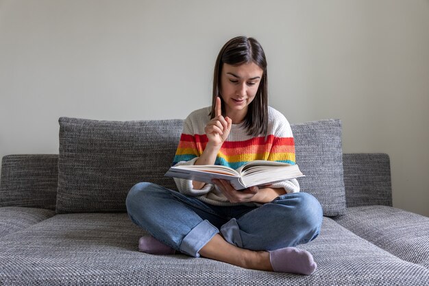Una ragazza con un maglione dai colori vivaci legge un libro sul divano di casa.