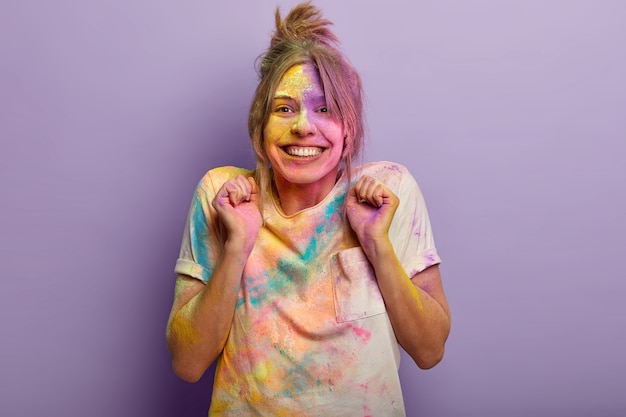 Una ragazza felice e ottimista stringe i pugni dalla gioia, ha pettinato i capelli, gioca con la polvere colorata, si rallegra della tradizionale festa di Holi e della primavera imminente, imbrattata di coloranti indiani. Concetto di vacanza